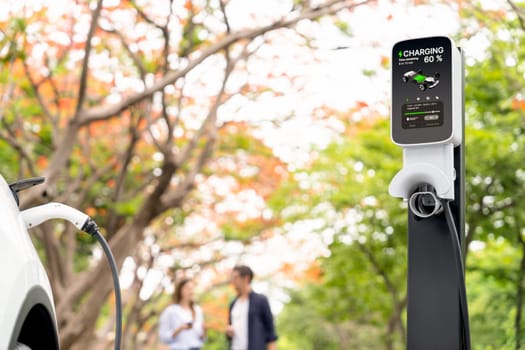 Focused EV car recharging battery on blurred background of lovey couple during autumnal road trip travel with electric vehicle recharging battery. Eco friendly travel on vacation during autumn. Exalt