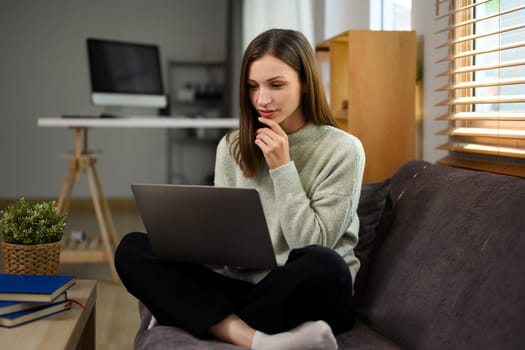 Attractive young woman in casual clothes suing laptop sitting on couch at home.