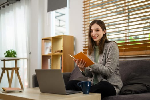 Pretty young woman holding book and using laptop on couch at home.