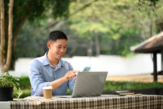 Smiling adult asian man using mobile phone and working with laptop at outdoor cafe.