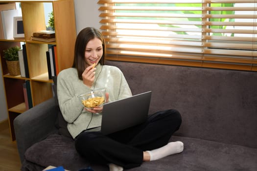Relaxed young caucasian woman eating popcorn and watching movie on a laptop at home.