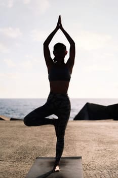 backlight of young woman in sportswear doing tree position on her yoga mat by the sea, concept of mental relaxation and healthy lifestyle