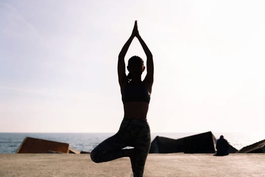 backlight of young woman in sportswear doing yoga tree position by the sea, concept of mental relaxation and healthy lifestyle