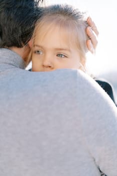 Little girl peeks out from behind her daddy shoulder while sitting in his arms. Back view. High quality photo