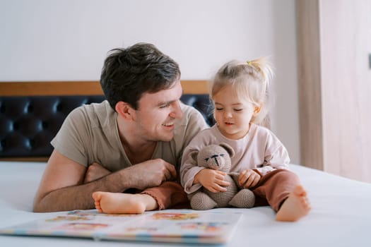 Smiling dad looking at little girl lying next to her in front of a book on the bed. High quality photo