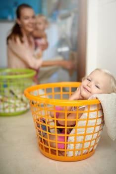 Happy baby, portrait and relax with mom in laundry basket for fun childhood, game or chore day at home. Young little girl with smile in bucket while mother washing clothing in machine for hygiene.