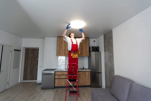 Young man installing ceiling lamp on stepladder in kitchen.