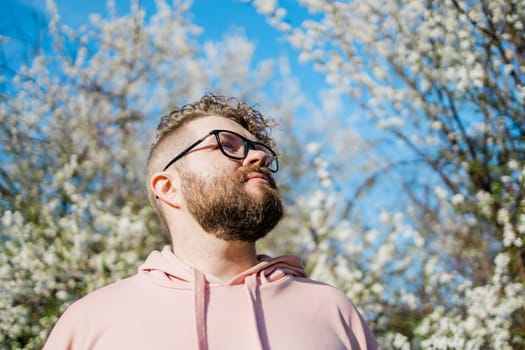 Male bearded guy standing under branches with flowers of blooming almond or cherry tree in spring garden. Spring blossom. Copy space