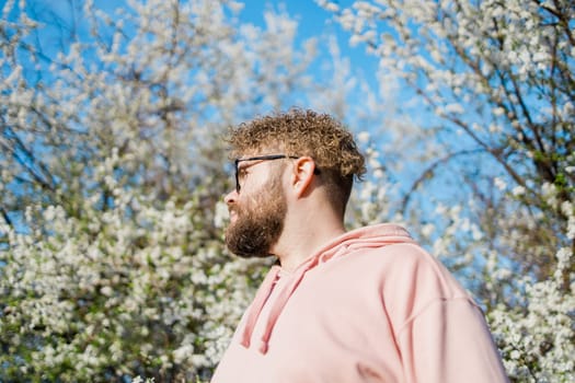 Male bearded guy standing under branches with flowers of blooming almond or cherry tree in spring garden. Spring blossom. Copy space