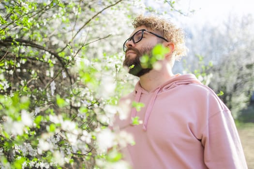 Male bearded guy standing under branches with flowers of blooming almond or cherry tree in spring garden. Spring blossom. Copy space