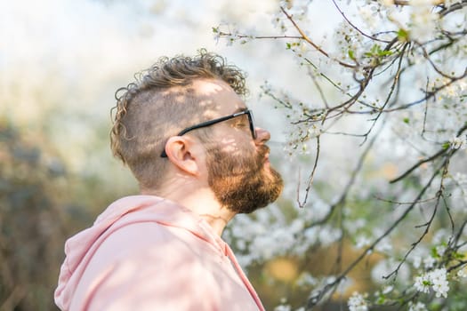 Male bearded guy standing under branches with flowers of blooming almond or cherry tree in spring garden. Spring blossom. Copy space