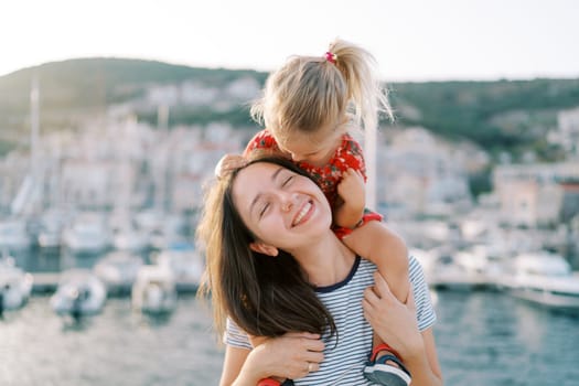 Little girl almost kisses her smiling mother on the cheek while sitting on her shoulders on the seashore. High quality photo