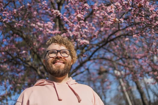 Man allergic enjoying after treatment from seasonal allergy at spring. Portrait of happy bearded man smiling in front of blossom tree at springtime. Spring blooming and allergy concept. Copy space.