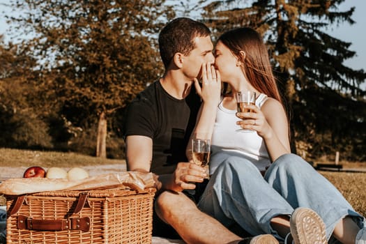 One beautiful caucasian young couple kissing covering their faces with their hands and holding glasses of champagne sit on a bedspread with a wicker basket and fruits in the park on a summer sunny day, close-up side view.