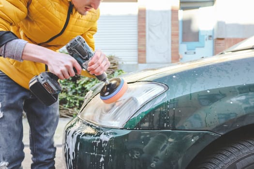 A young caucasian guy in a yellow jacket is drilling a drill with a disk with a sponge and polishing the headlight of his car with detergent soap on the city street in front of the house, close-up side view.Concept home car wash and cleaning with polishing car headlights.