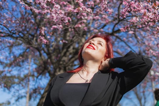 Red haired woman wearing stylish outfit near blossoming sakura in park. Fashionable spring look. Springtime female portrait