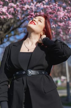 Red haired woman wearing stylish outfit near blossoming sakura in park. Fashionable spring look. Springtime female portrait