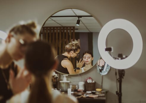 One young handsome Caucasian makeup artist applies protective cream to the face of a girl sitting in a chair, reflected in a round wall mirror early in the morning in a beauty salon, close-up side view. Step by step.