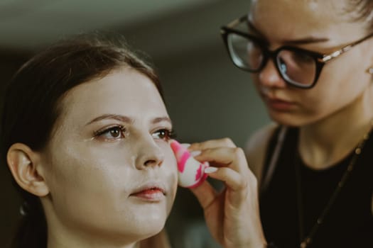 One young handsome Caucasian makeup artist applies foundation with a pink sponge to the cheekbones of the face of a girl sitting in a chair early in the morning in a beauty salon, close-up side view. Step by step.
