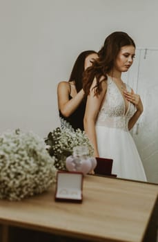 Portrait of a beautiful brunette bride and bridesmaid who fastens the clasp of a necklace around her neck, standing in a room reflected in a mirror, close-up side view.