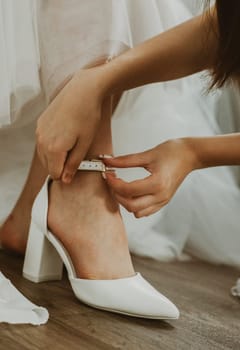 The hand of an unrecognizable bridesmaid helps to put white wedding shoes on the bride's leg, close-up side view.