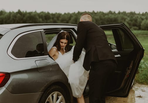 One young handsome Caucasian groom in a black suit stands leaning from the back and helps the bride get out of the car holding the hem of her dress, preparing for the wedding on a cloudy rainy day, side view close-up.