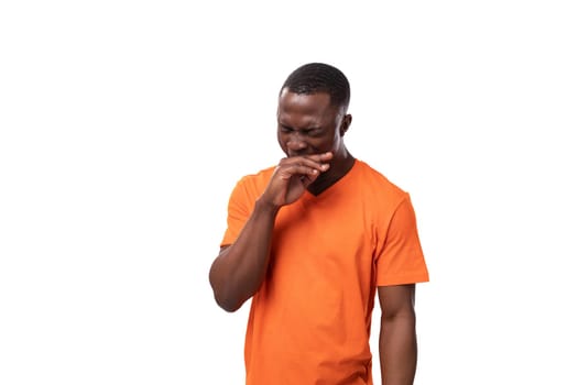 young african guy dressed in an orange t-shirt shyly covered his mouth with his hand on a white background with copy space.