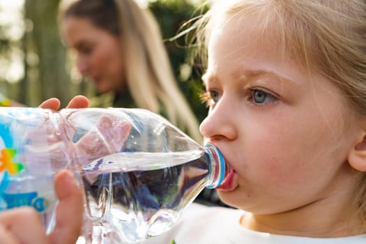 Little blonde girl drinking clean water from bottle in park close up