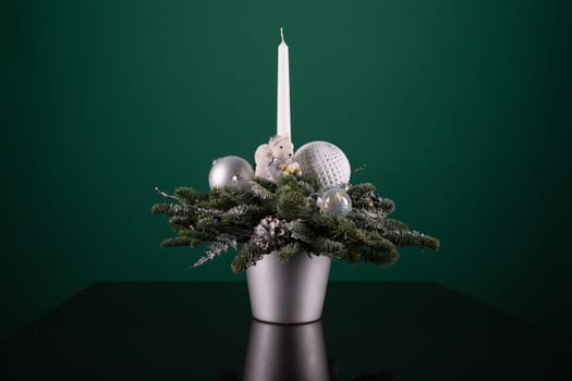 A silver vase sits on a table, filled with festive Christmas decorations like baubles and garlands. Next to it, a white candle burns softly, casting a warm glow on the scene.