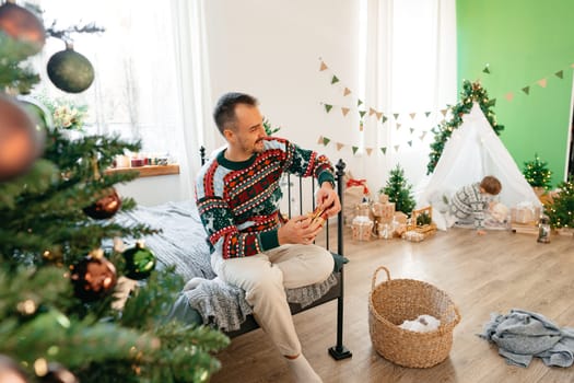 Young man in sweater sits on bed near Christmas tree at home