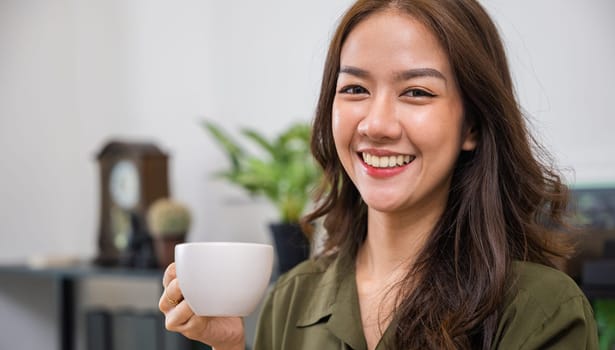 Portrait of Happy beautiful woman rest in summer after wakeup at office room, Asian young woman hands holding hot coffee or tea cup for drink in morning while posing, white porcelain mug mock up