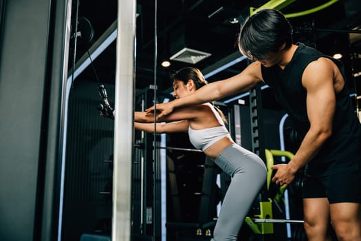 Hispanic man supporting and guiding a beautiful Asian woman through her pull down cable machine workout at the fitness center. GYM healthy lifestyle concept