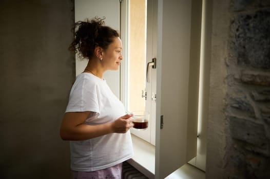 Attractive young adult woman in pajamas standing by the window with a cup of hot drink, admiring the beautiful view from the window.