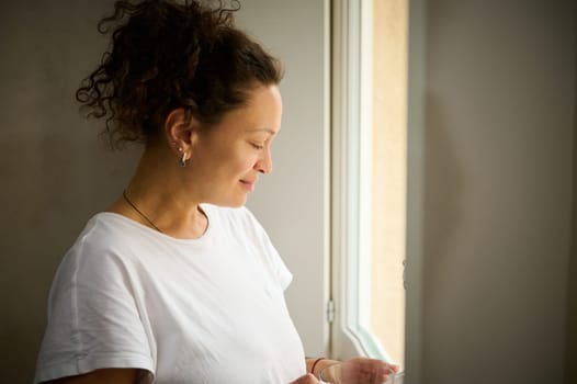 Close-up side portrait of a multi ethnic curly haired woman holding a cup of hot drink, smiling admiring the view from her window