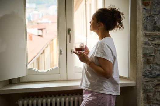 Authentic portrait of beautiful young woman admiring the beautiful view of a medieval city in Italy, standing by the window in the hotel room with cup of hot drink. People. Travel. Lifestyle