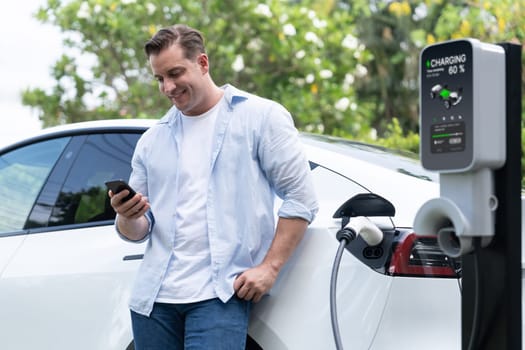 Modern eco-friendly man recharging electric vehicle from EV charging station, using Innovative EV technology utilization for tracking energy usage to optimize battery charging on smartphone.Synchronos