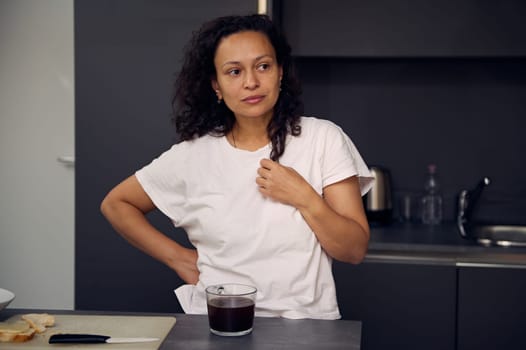 Pensive young woman in white pajamas t-shirt, standing at kitchen table with a cup of freshly brewed coffee drink, dreamily looking away
