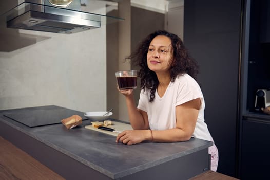 Curly haired brunette holding a cup of coffee, smiling and dreamily thoughtfully looking away, standing at kitchen counter while taking her breakfast in the gray minimalist home kitchen interior