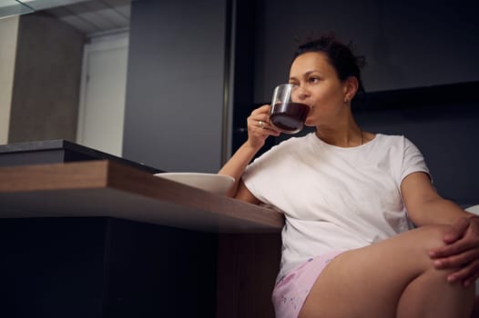 View from the bottom to a relaxed woman in stylish pajamas, sitting at kitchen table and drinking coffee during her breakfast