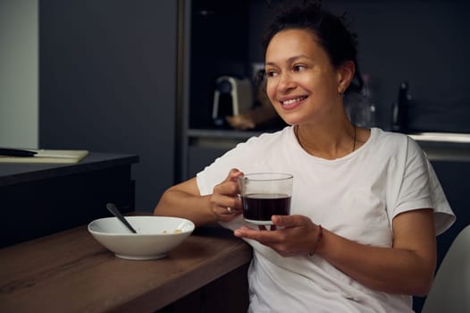Portrait of a joyful young woman enjoying a cup of coffee at home. Attractive pretty lady drinking hot freshly brewed coffee, smiling looking away, sitting at kitchen table while taking a breakfast