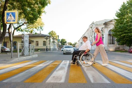 Young female caregiver pushing wheelchair with mature female person with disability across city street