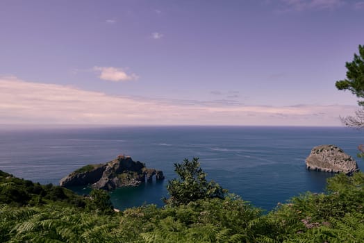 Hermitage of San Juan de Gaztelugatxe on a sunny day. calm sea, rocky formation, holes, frame of branches and trees