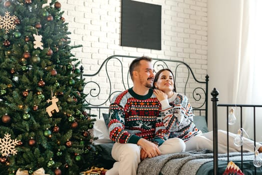Young woman and man sitting on bed in sweaters and hugging near Christmas tree close up