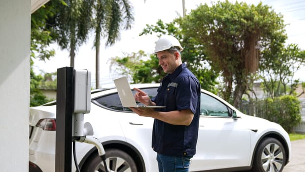 Qualified technician working on home EV charging station installation, making troubleshooting and configuration setup on charging system with laptop for EV at home. Synchronos