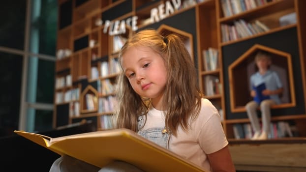 Young smart caucasian girl picking reading a book while sitting at library. Clever child learning, studying, open a books at library. Attractive kid turning page with blurring background. Erudition.