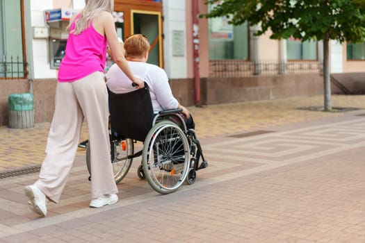 Back view of young woman helping mature woman in wheelchair strolling in the city
