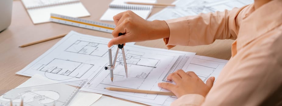 Professional architect drawing blueprint by using divider on the table with stationary and architectural document scatter around at architectural office. Closeup. Focus on hand. Delineation.