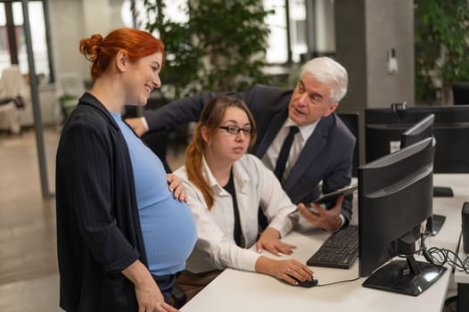 An elderly man, a Caucasian woman and a pregnant woman are discussing work issues at the computer