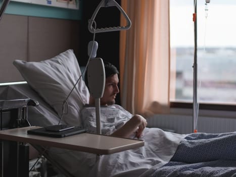 A young guy patient with sad emotions in uniform holds his hand to his stomach in pain after surgery in bed under a dropper in front of a window in a hospital, close-up side view.Health care concept.
