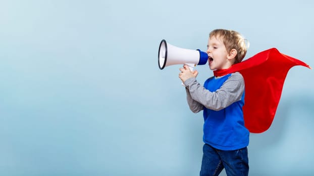 Boy with red cape shouting into megaphone, blue background. Studio child portrait. Leadership and communication concept. Design for banner, poster, card.